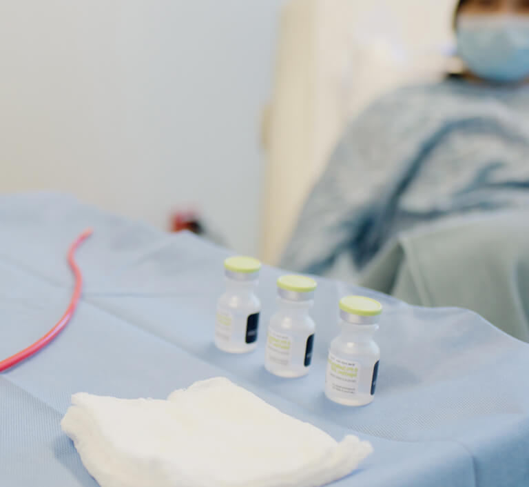 Medical preparation setup in a clinical setting: three small glass vials with green caps and labels (containing BCG and ANKTIVA) placed on a sterile blue drape next to a red catheter tubing, white gauze pad, and blurred healthcare professional in mask and gown in the background