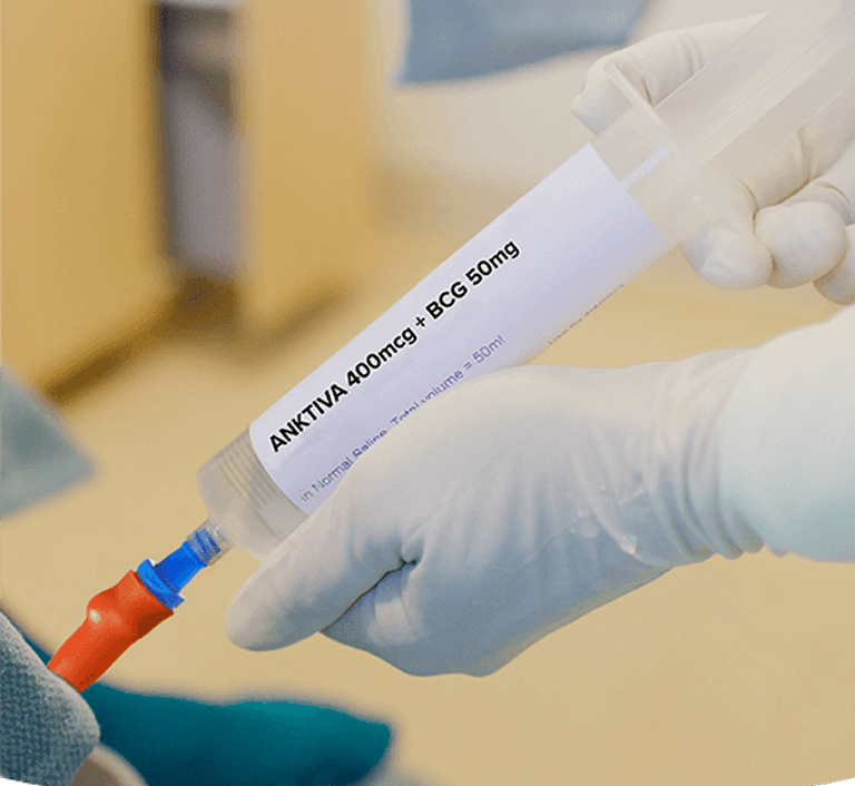 Gloved hands holding a syringe labeled "ANKTIVA 400mcg + BCG 50mg" connected to a catheter, in a blurred clinical setting